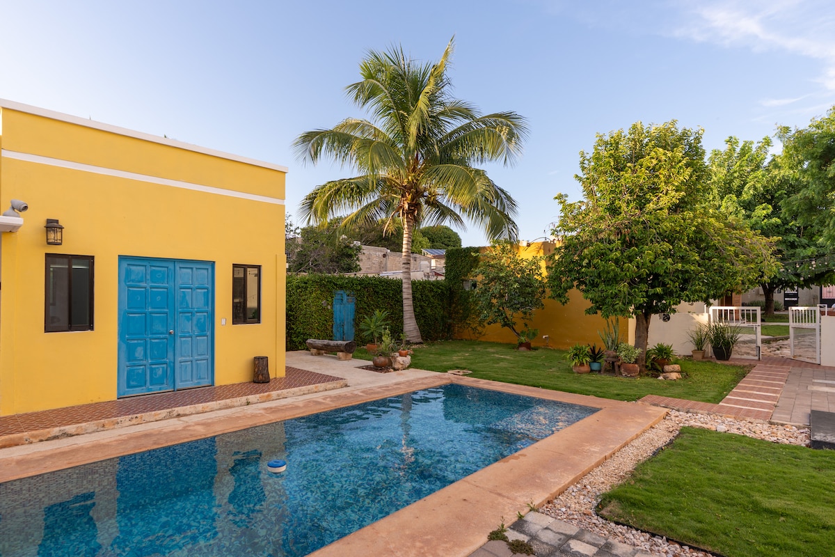 A vibrant yellow building is seen adjacent to a clear swimming pool, surrounded by greenery. Towering palm trees and shrubs frame the area. A neatly maintained garden features a stone pathway and potted plants, creating an inviting outdoor space for relaxation.