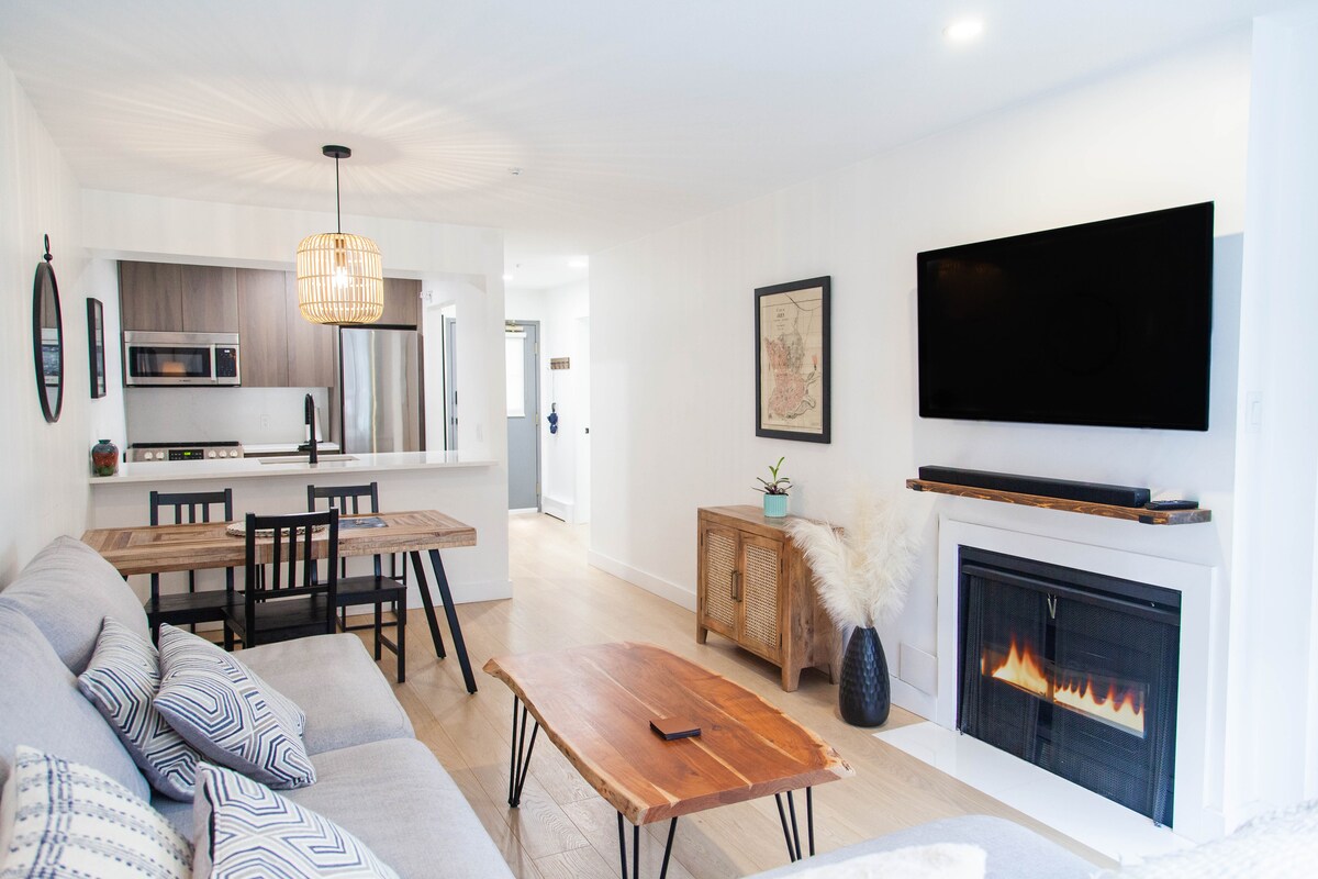 An open-concept living space showcases a comfortable gray sofa and a rustic wooden coffee table. A wall-mounted television is positioned above a modern fireplace. The kitchen area, featuring sleek cabinetry and appliances, is subtly visible in the background, enhancing the home's welcoming feel.