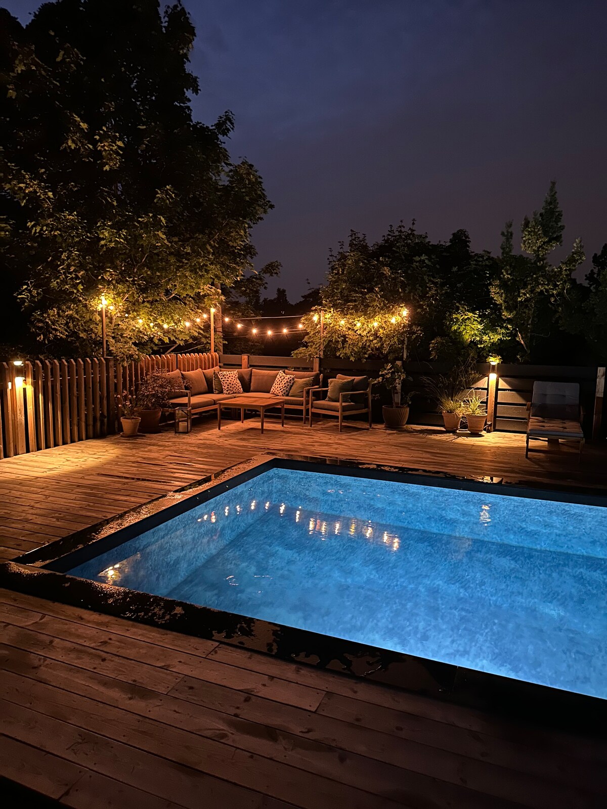 A serene outdoor space is illuminated by warm string lights, highlighting a wooden deck surrounding a clear pool. Comfortable seating is arranged nearby, offering a cozy area for relaxation amidst the lush greenery.
