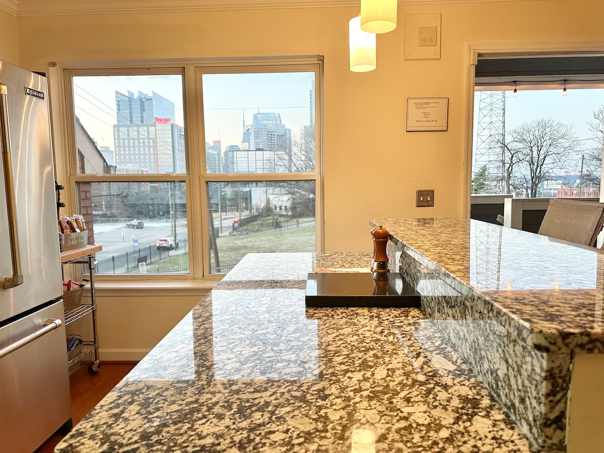 A spacious kitchen showcases granite countertops and a central island under modern pendant lighting. Large windows offer a clear view of the downtown skyline, while stainless steel appliances and subtle decor enhance the kitchen's functionality and style.