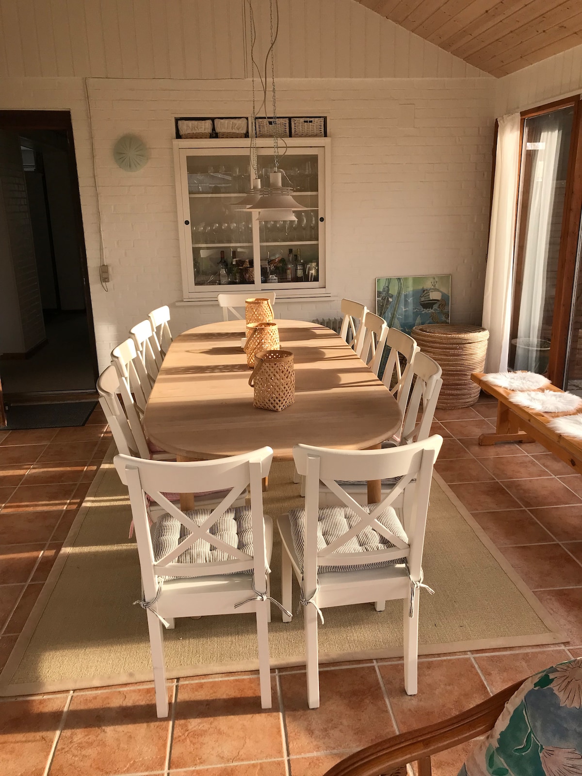 A spacious dining area features a long wooden table surrounded by ten white chairs. The room is filled with natural light, highlighting cozy decorative elements. A light fixture hangs above the table, complemented by warm tones of the tiled floor.