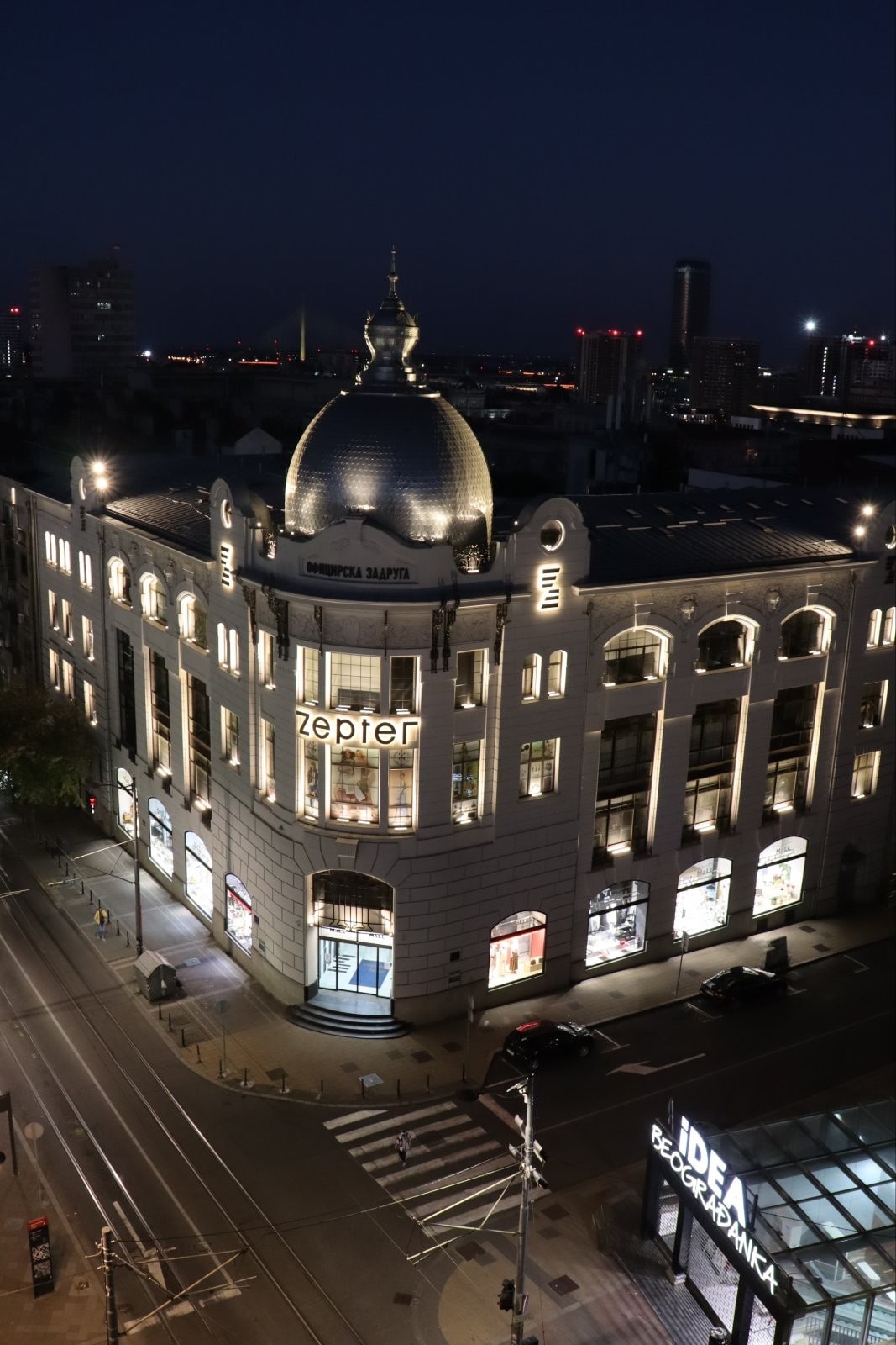 The image captures a splendid building at night, illuminated by soft lighting. The architectural details are showcased, including a prominent dome and large windows. The surrounding area features streets and city lights, conveying a sense of vibrancy and urban charm.