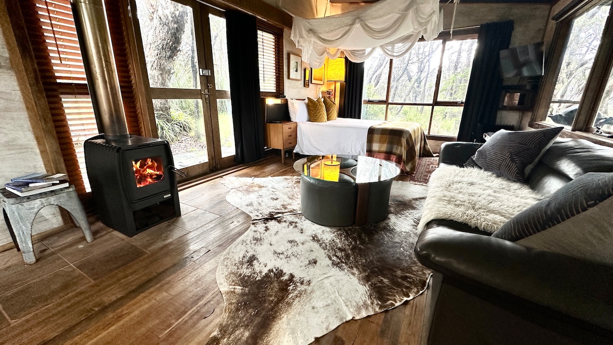 A warm, inviting interior is showcased with a cozy seating area featuring a leather couch. A round coffee table sits atop a soft, textured area rug. A wood heater provides additional warmth next to the large windows, which offer views of the surrounding bushland.