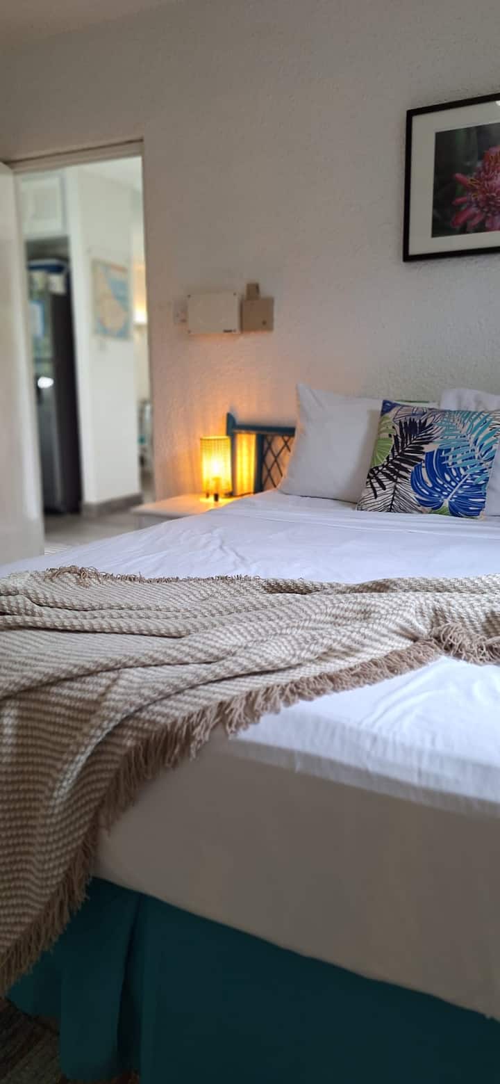 Unwind in our air-conditioned primary bedroom, featuring a queen bed, crisp white linens, tropical accents, and warm lighting that create the perfect soothing sanctuary after a day in the sun.