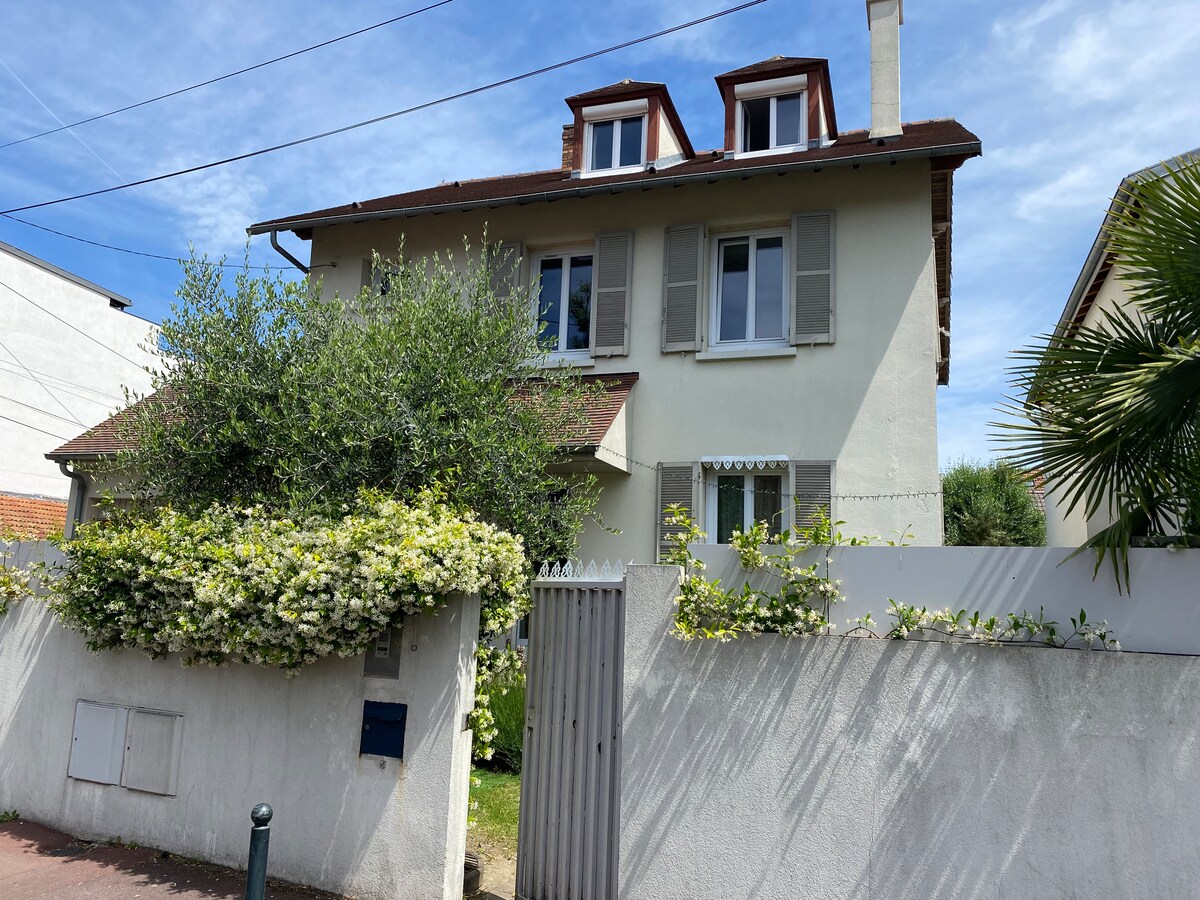 The exterior of a two-story house is visible, featuring a light-colored facade with white shutters and a brown roof. A lush green hedge and flowering plants create a welcoming entrance, while a fenced yard adds privacy. Clear blue skies enhance the home's inviting appearance.
