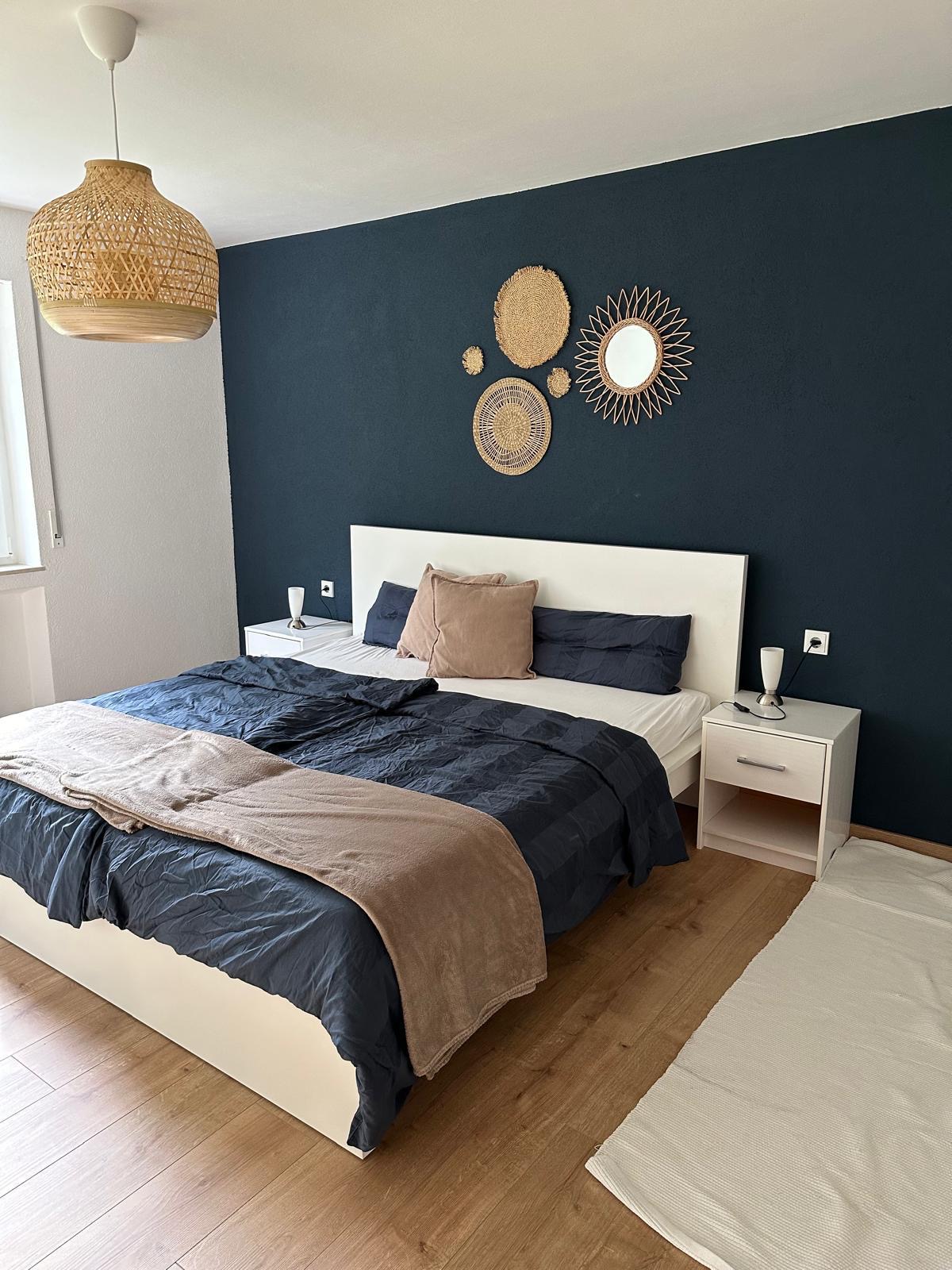 A serene sleeping area features a spacious bed with neutral bedding and decorative pillows. A bedside table with glassware is positioned on either side, and a decorative mirror and wall art add interest to the dark accent wall.
