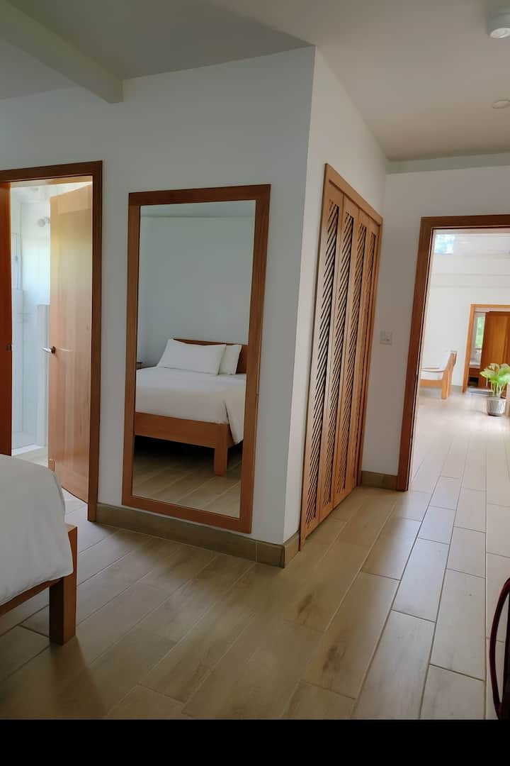 Bright, minimalist bedroom with warm cedar finishes, full-length mirror and neutral tones. Large window with light curtains fills the space with natural light, creating a calm and welcoming atmosphere.