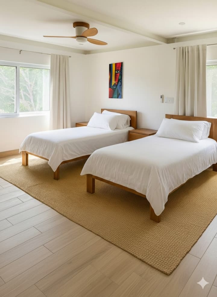Bright twin bedroom with two wooden beds, soft neutral tones and large windows overlooking greenery. Includes modern ceiling fan, air-conditioning unit, desk and potted plant for a calm, natural feel.