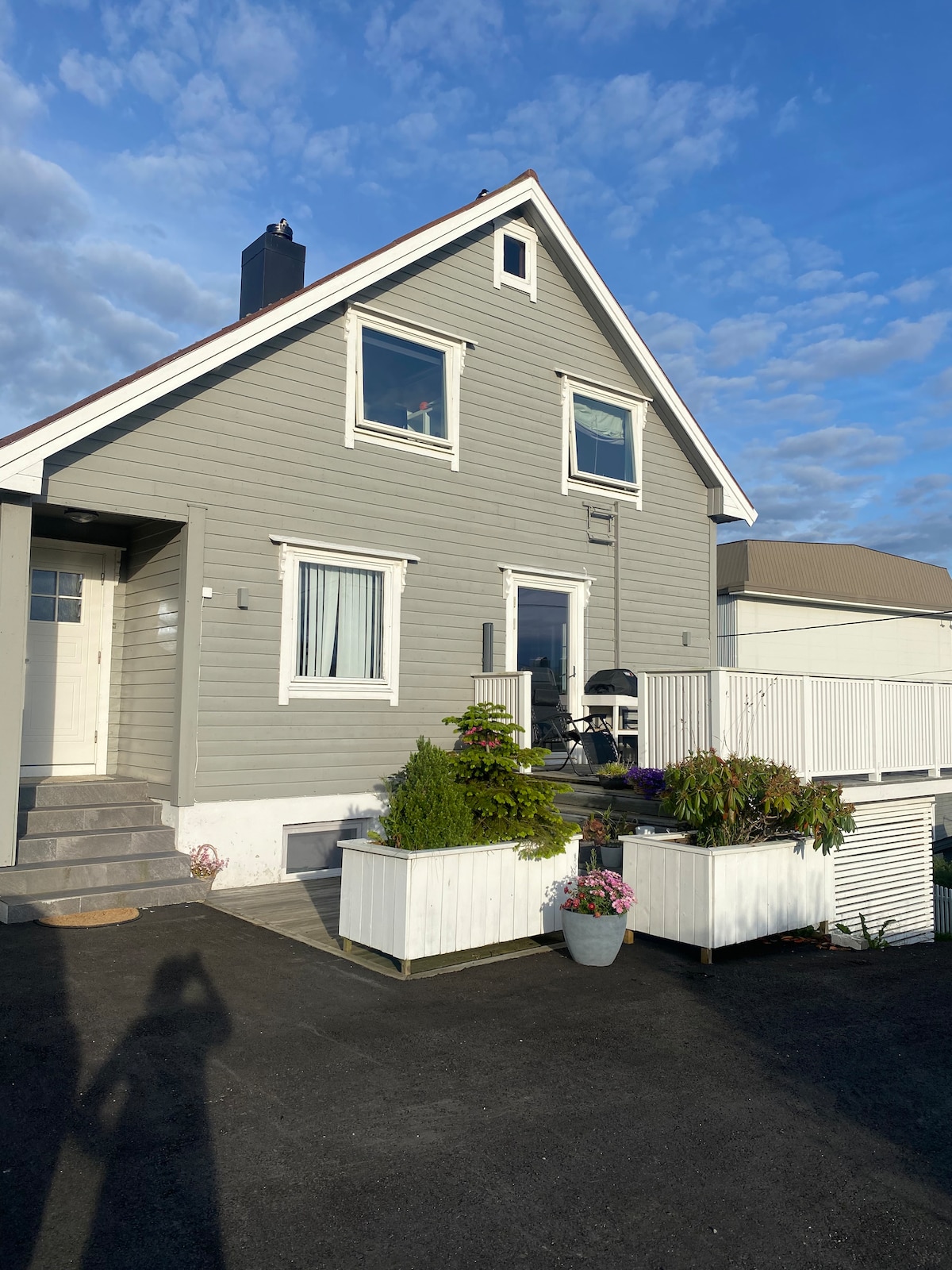 The exterior of a two-story house is depicted, featuring a light grey facade and white trim. Several windows are visible, and colorful flower boxes add interest to the porch area. A small set of stairs leads to the entrance, with a clear sky in the background.