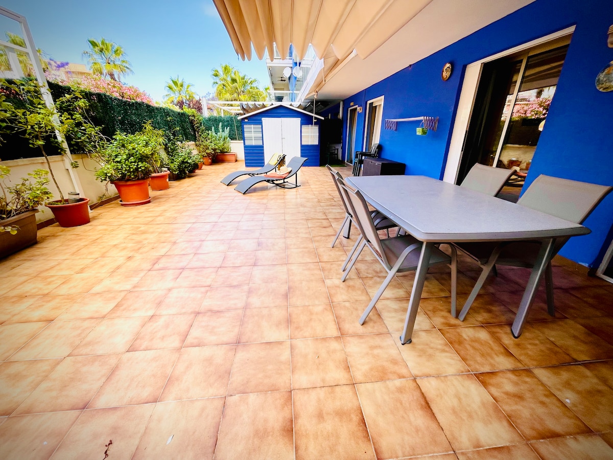 A spacious terrace is shown with tiled flooring and painted blue walls in the background. A large dining table and chairs are situated alongside a sun lounger, surrounded by various potted plants. The area is bright and open, offering plenty of sunlight.