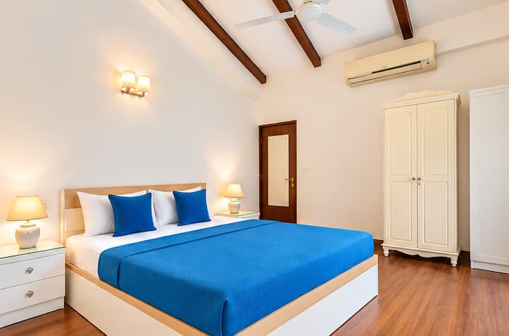 A bright and airy bedroom with a comfortable bed, crisp white linens, vibrant blue accents, warm wooden flooring, and beautiful greenery views from the balcony.