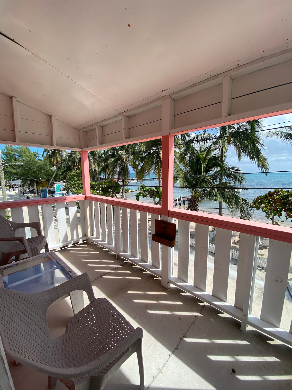 A charming balcony space is shown, featuring two white chairs and a small glass table. Lush palm trees and the Caribbean Sea are visible through the open railings, allowing natural light to fill the area. The coastal view creates a relaxing ambiance.