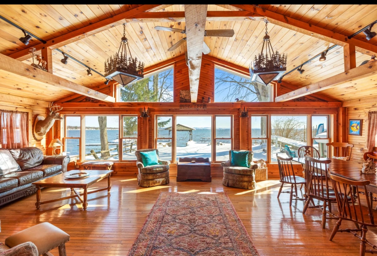 A spacious living area features high wooden ceilings, with large windows offering views of the lake. Comfortable seating is arranged around a coffee table, complemented by a patterned area rug. Natural light fills the room, creating a warm and inviting ambiance.