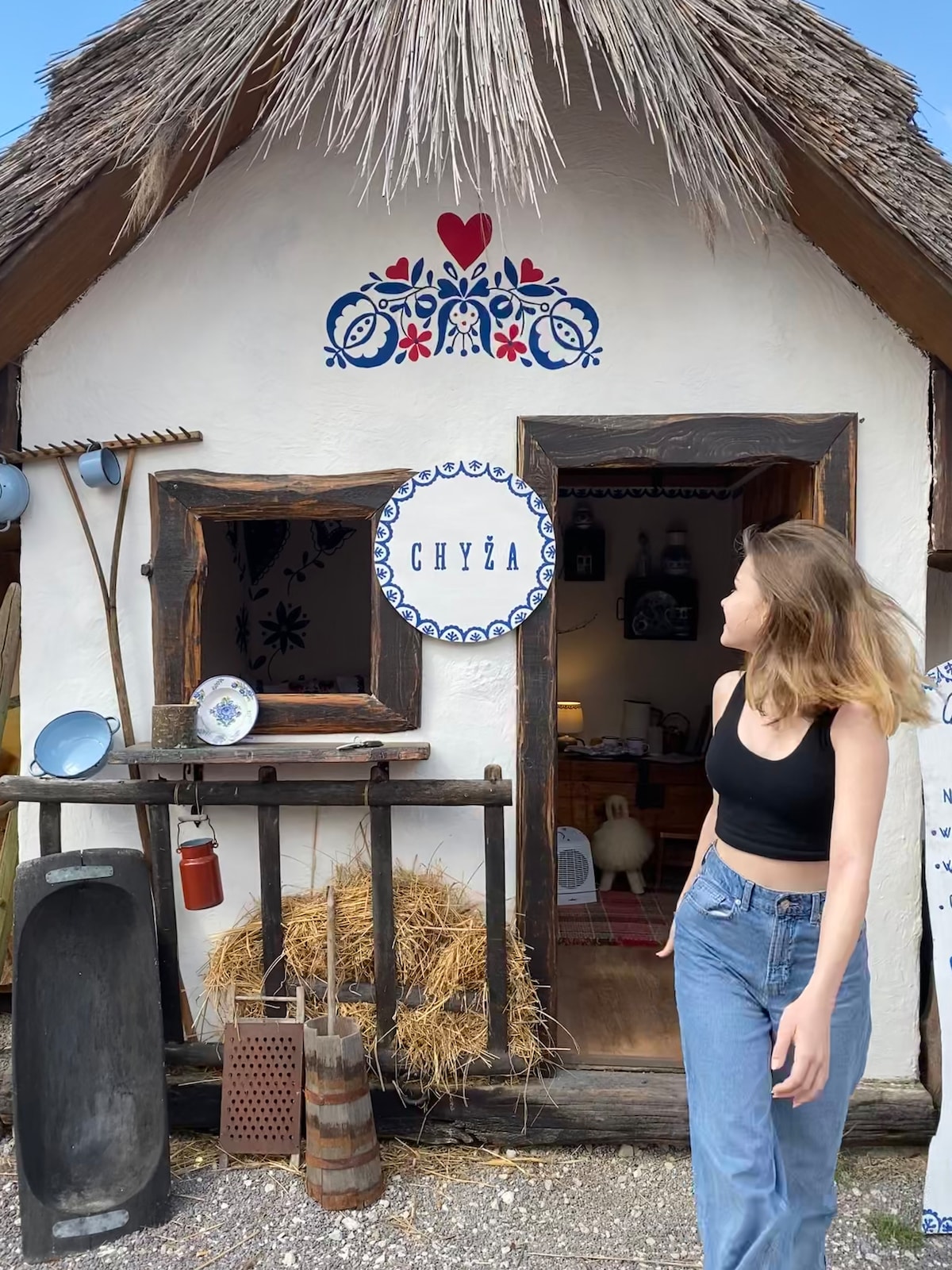 The entrance to a traditional folk hut is displayed, featuring a thatched roof and decorative painting above the door. Straw is seen arranged at the base, with rustic tools and pottery positioned nearby, showcasing the cultural essence of the space.