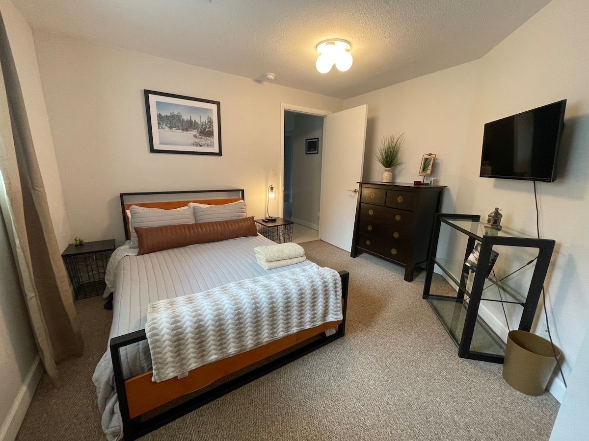 A cozy bedroom is featured, furnished with a spacious bed dressed in soft linens. A wooden dresser and a sleek glass side table offer storage options. Natural light filters through the window, complemented by a framed landscape photograph on the wall.