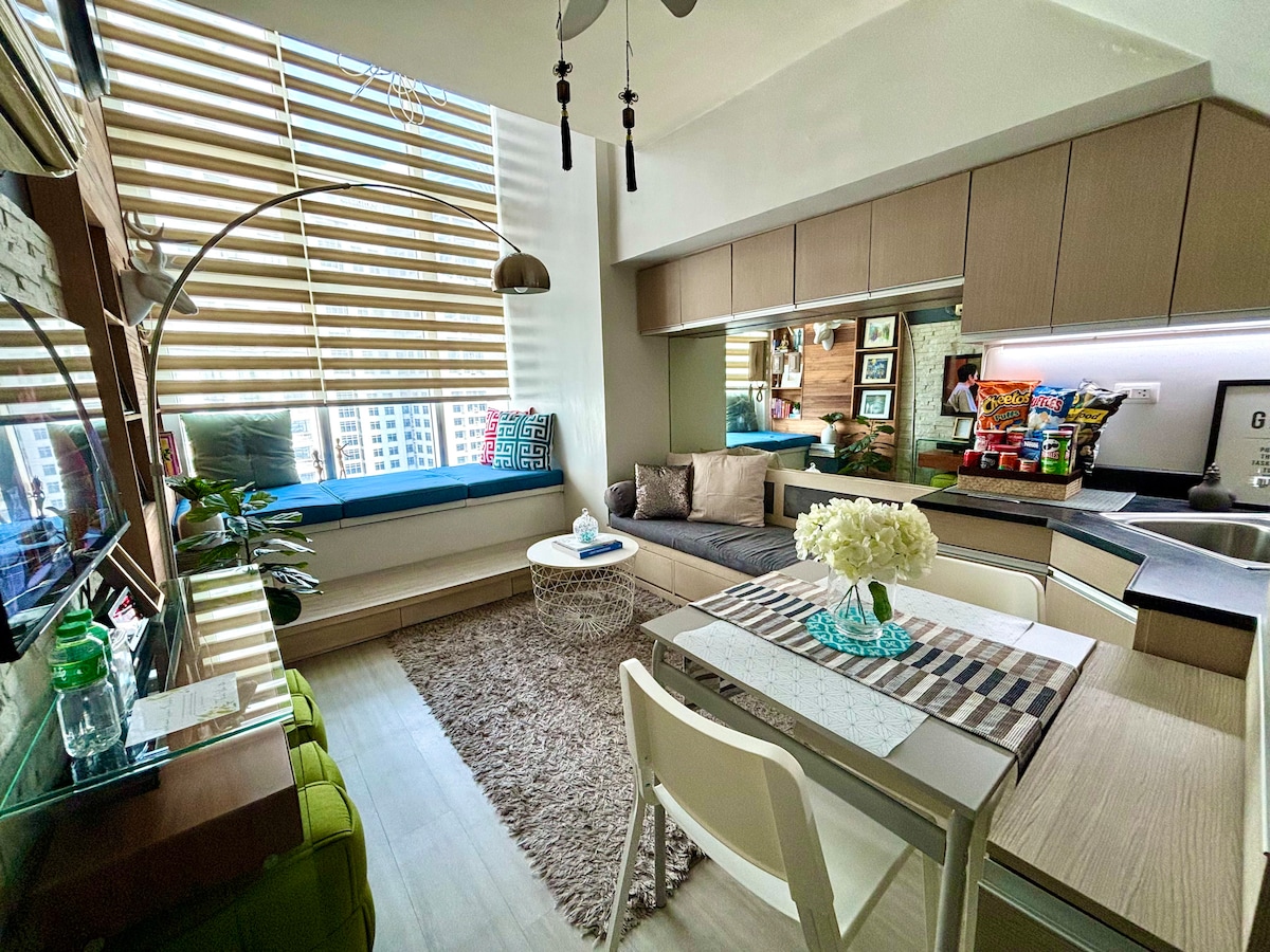A modern loft features a dining area with a round table and stylish chairs. A cozy seating nook is accented by cushions and a coffee table, while a well-equipped kitchenette is visible. Natural light streams through large windows adorned with sleek blinds, creating a bright atmosphere.