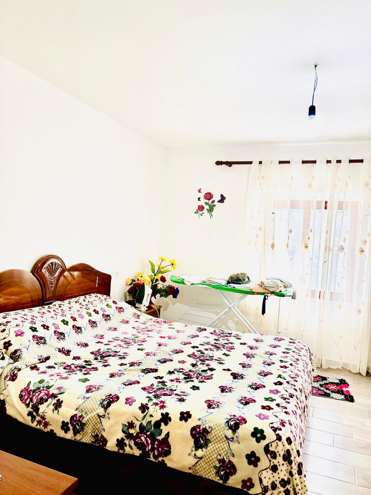 A bright bedroom features a spacious bed covered with a floral-patterned blanket. Natural light filters through sheer curtains next to the window. An ironing board and fresh flowers add a touch of practicality and charm to the serene space.
