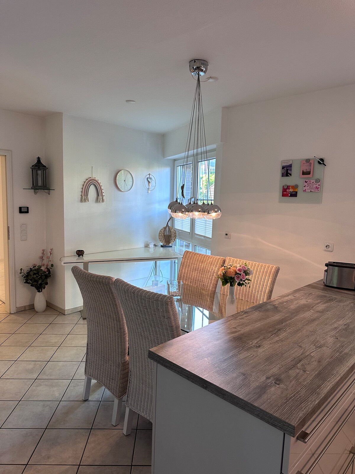 A well-lit dining area features a round glass table surrounded by four wicker chairs. Natural light floods through the large windows, highlighting the minimalist decor on the walls. A modern pendant lamp hangs gracefully above the table, enhancing the open and airy atmosphere.