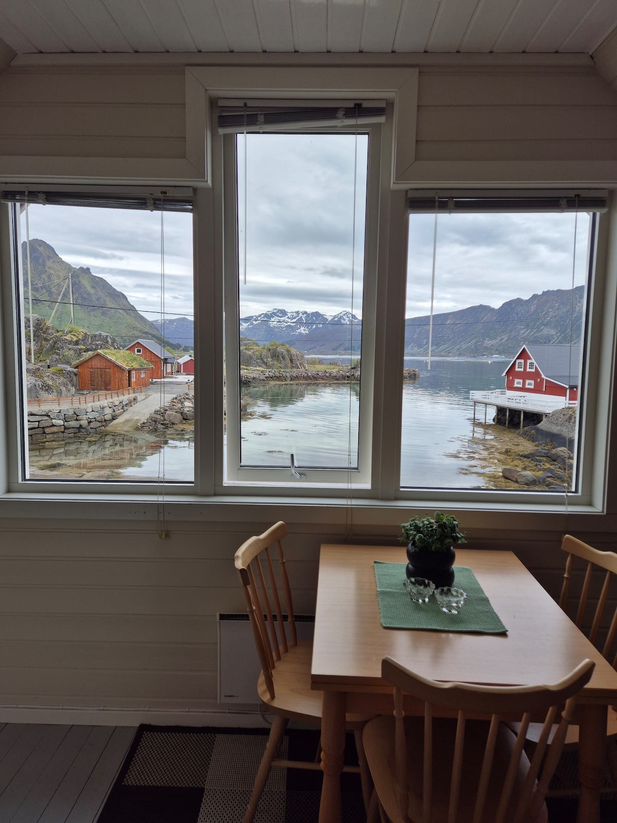 Large windows frame a serene view of the surrounding mountains and calm waters. A small wooden table is set with a green placemat and a decorative plant. The presence of traditional red buildings is visible across the water, enhancing the natural setting.