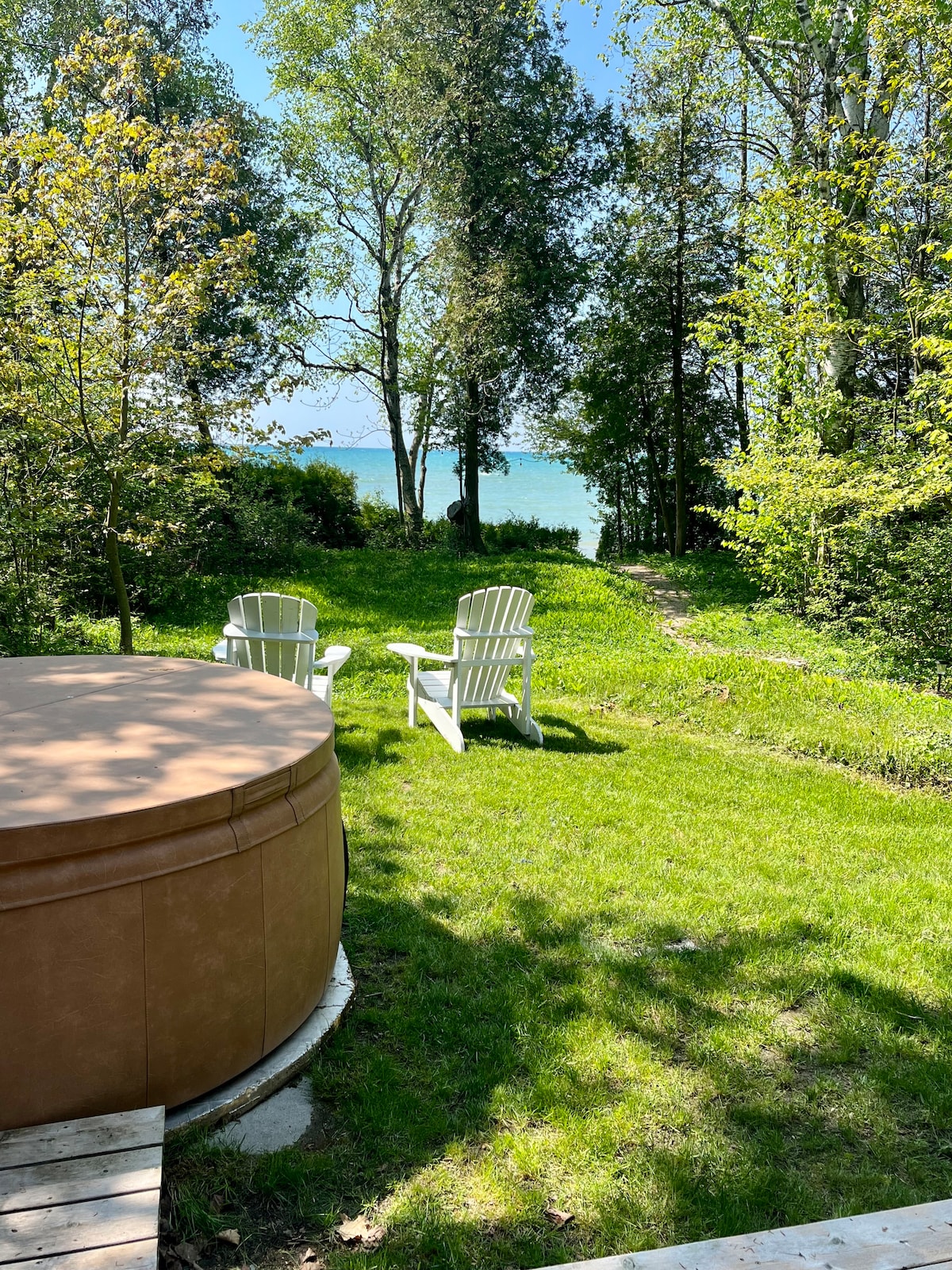 Two white Adirondack chairs are positioned on lush green grass, facing the serene waters of Lake Huron. A hot tub is situated nearby, with trees providing a natural backdrop. The clear blue sky and gentle sunlight enhance the tranquil outdoor setting.