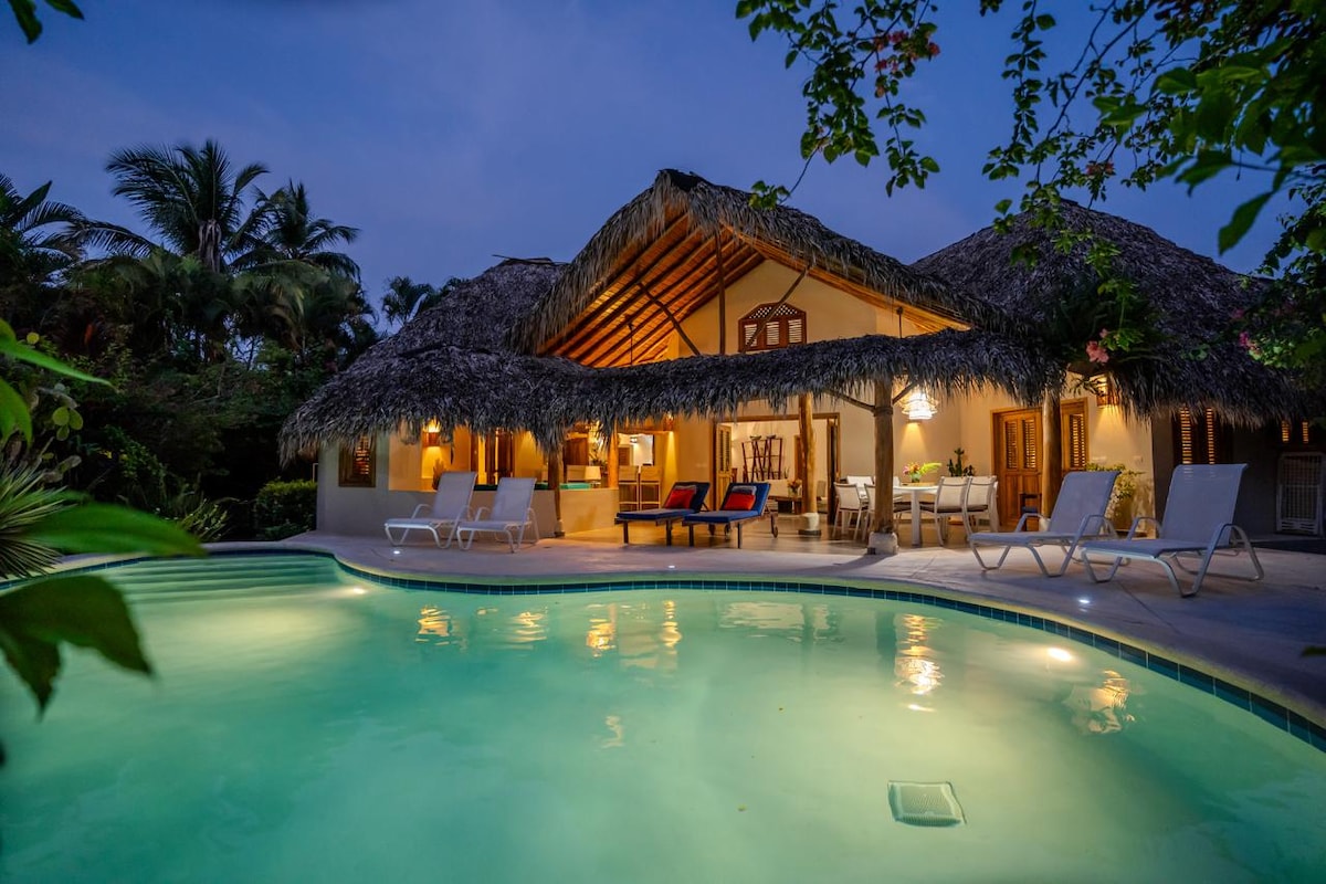 The villa is framed by lush tropical plants and features a thatched roof design. A sparkling pool reflects the evening sky, while lounge chairs are positioned nearby. The spacious covered terrace showcases seating arrangements and a dining area, inviting relaxation and social gatherings.