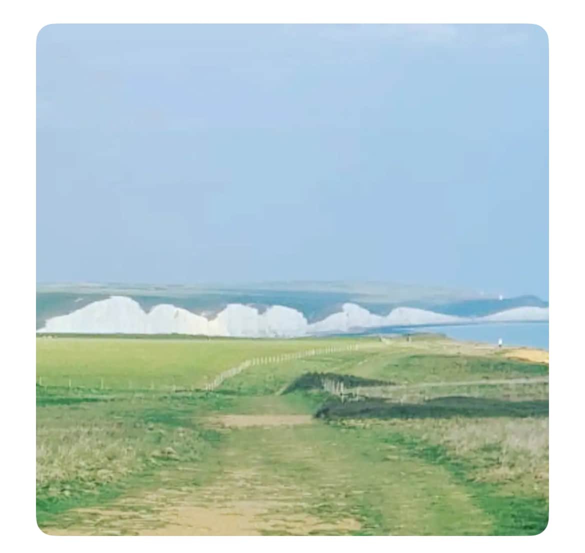 A scenic view captures a pathway leading through lush green fields, with white chalk cliffs visible in the distance. The coastline meets a calm sea under a clear sky, creating a serene landscape highlighted by gentle hues of green and blue.