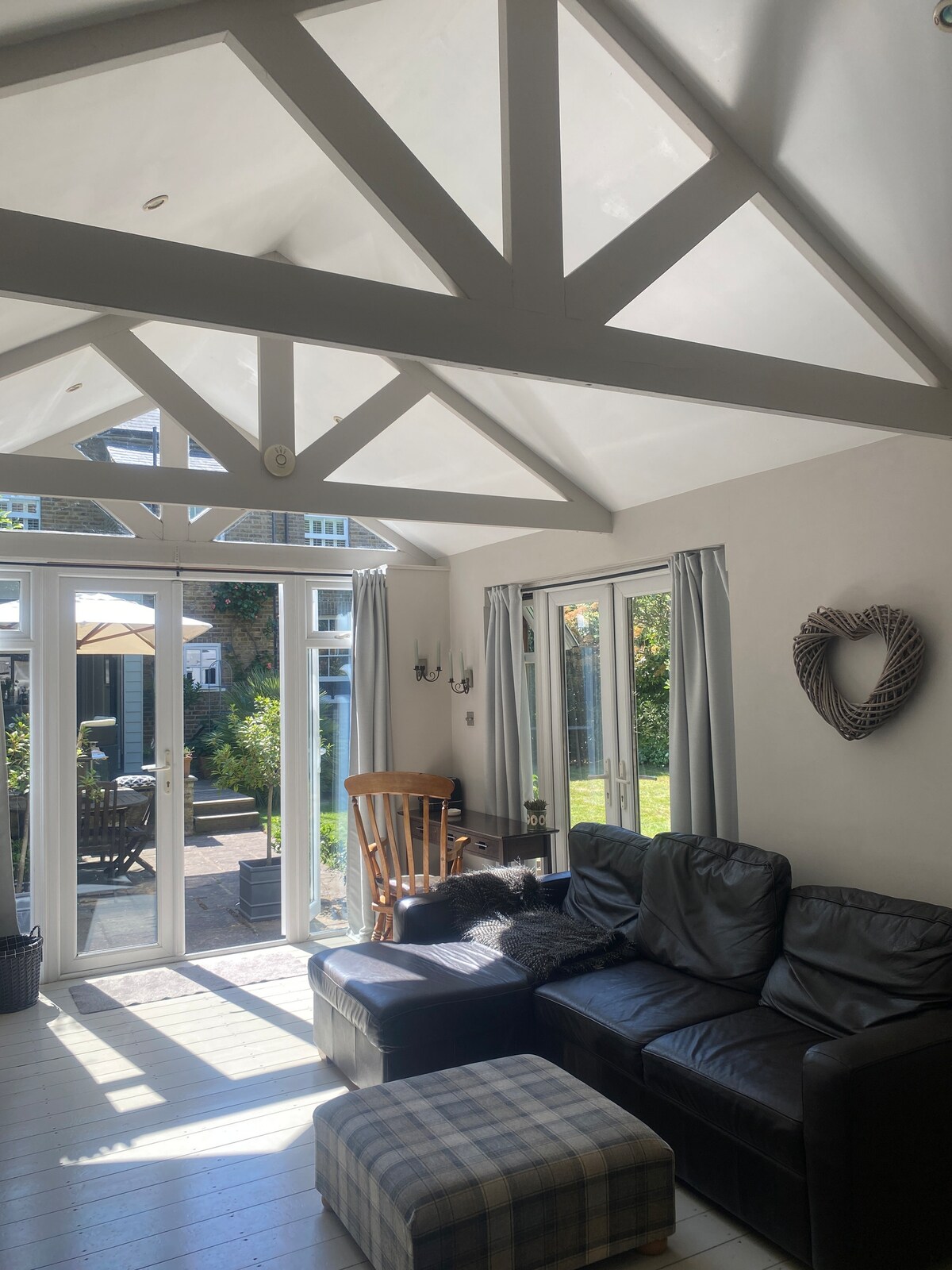 A bright living area is enhanced by high ceilings with exposed beams, featuring large, double glass doors that open to the garden. An L-shaped leather sofa, a checkered ottoman, and a wooden chair are arranged for comfort, while natural light streams in through the windows.