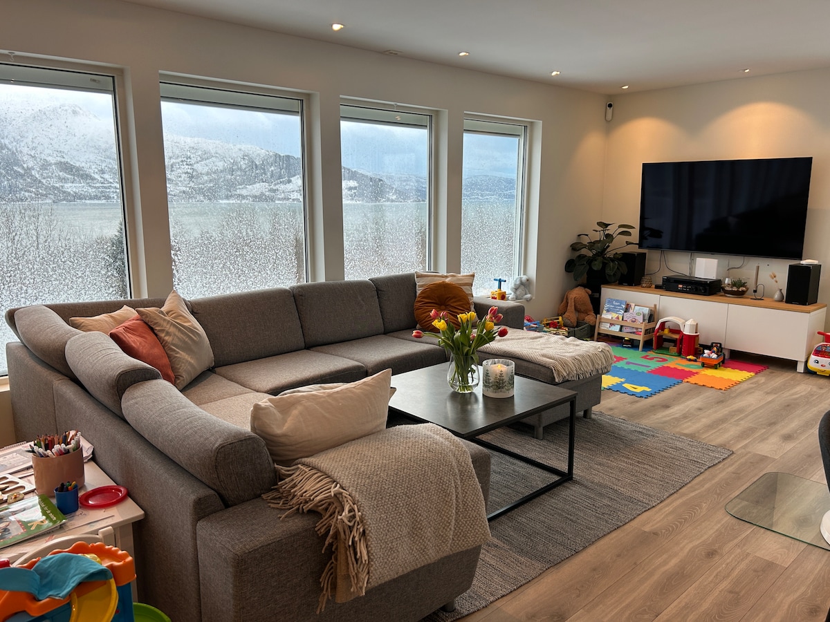 A spacious living area features large windows that offer views of the surrounding landscape, which appears soft-focused due to condensation. A sectional sofa is paired with a modern coffee table, while decorative pillows and a throw blanket add comfort. A TV is mounted on the wall, accompanied by children's toys and a play area.