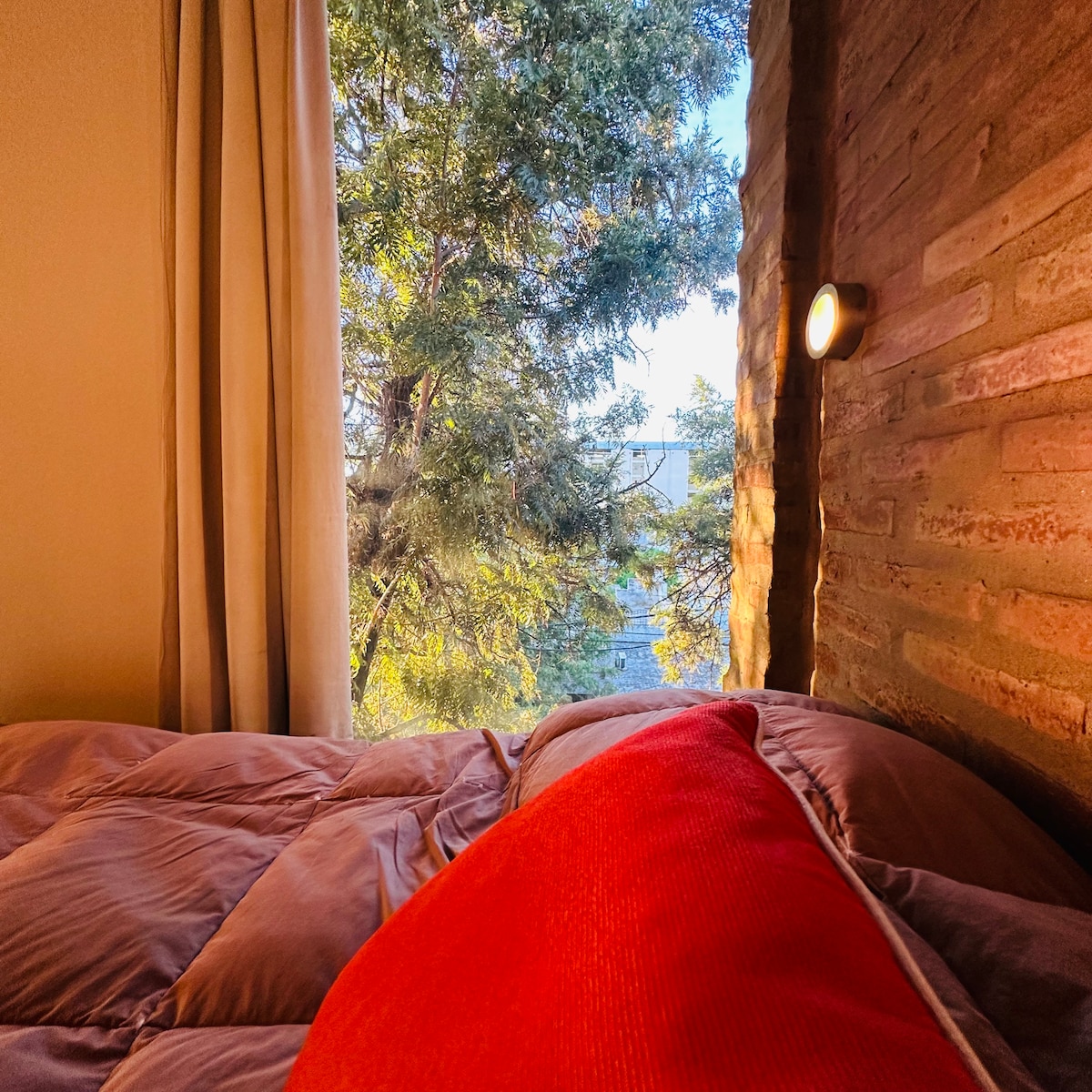 A comfortable bed with a plush, vibrant red pillow is positioned near a window. Soft curtains frame the view of lush green trees outside, allowing natural light to brighten the space. The exposed brick wall adds a rustic touch to the cozy setting.
