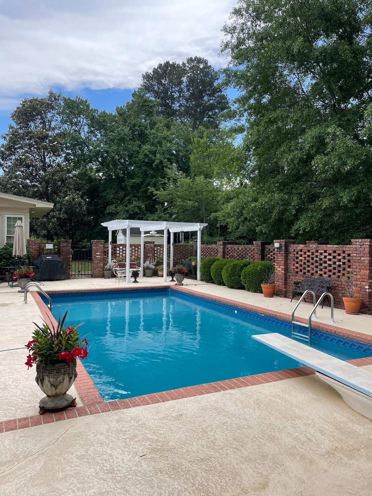 A well-maintained pool area is surrounded by brick walls and greenery. Lounge chairs are positioned near the water, while flower pots add pops of color. A shaded seating area with a gazebo provides a tranquil space for relaxation.