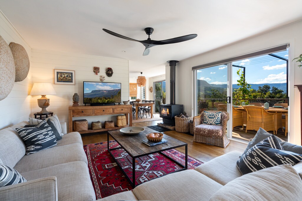 A cozy living area features a spacious sectional sofa facing a rustic wooden coffee table. A modern ceiling fan is visible above. A wall-mounted smart TV is positioned next to a wood-burning fire, with large glass doors opening to an outdoor seating area with scenic views.