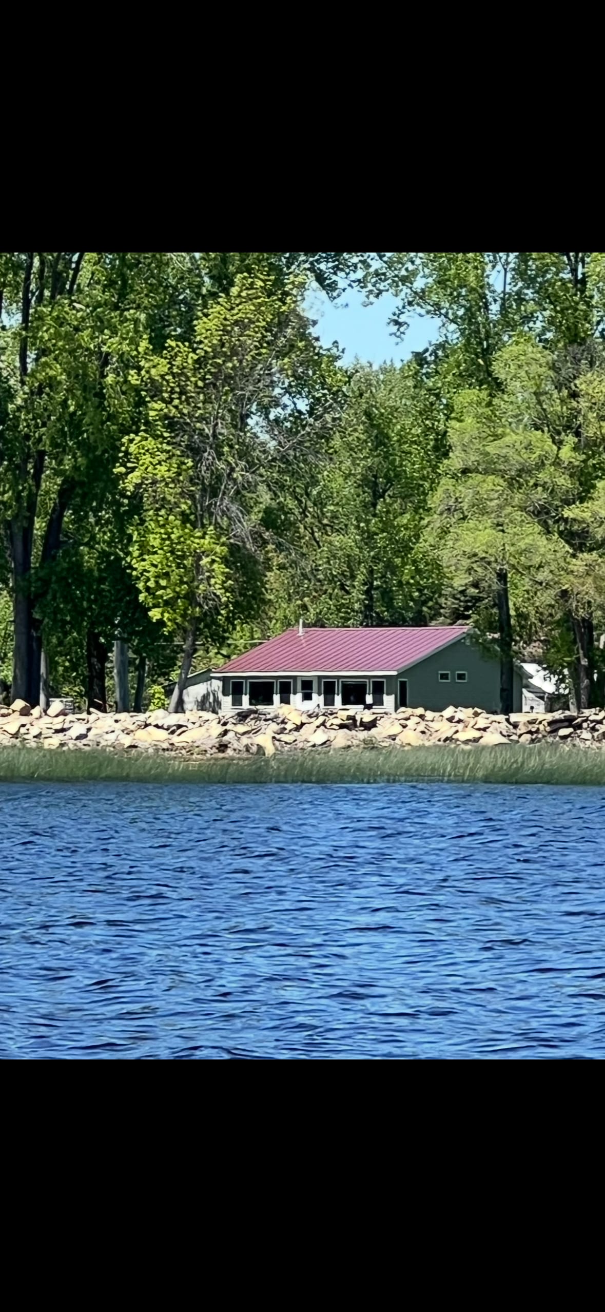 A charming waterfront home is situated among lush trees, featuring a red roof and large windows that reflect the tranquil setting. The exterior blends seamlessly with the natural landscape, while a stone shoreline frames the property's peaceful atmosphere.