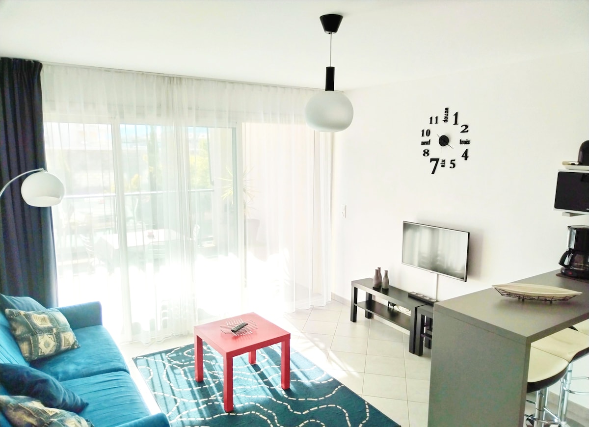 A living area is arranged with a blue sofa and a red coffee table placed on a patterned rug. A television sits on a stand, with a coffee maker nearby. Large glass doors allow natural light to fill the space, leading to a terrace outside.