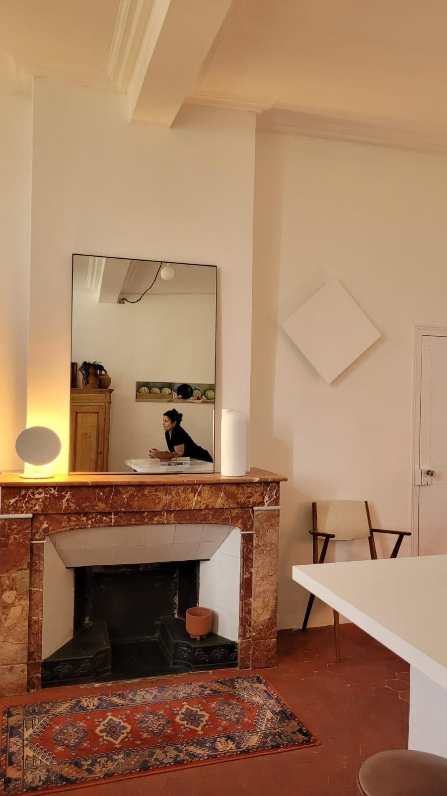 A fireplace made of patterned marble features a large mirror above it, reflecting the room's ambiance. A light emanates from nearby, creating a soft glow. Simple wooden furniture is positioned around the space, with a colorful rug adding warmth to the floor.