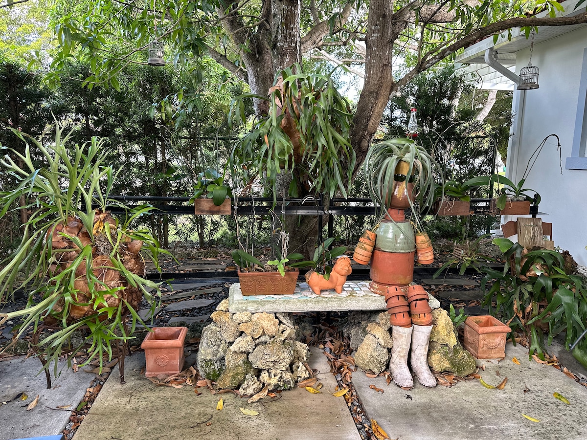 A serene outdoor area features a stone path leading to a rustic bench surrounded by various plants. Decorative terracotta figures and potted plants add charm, while a backdrop of lush greenery creates a peaceful atmosphere. Natural light filters through the trees, enhancing the tranquility.