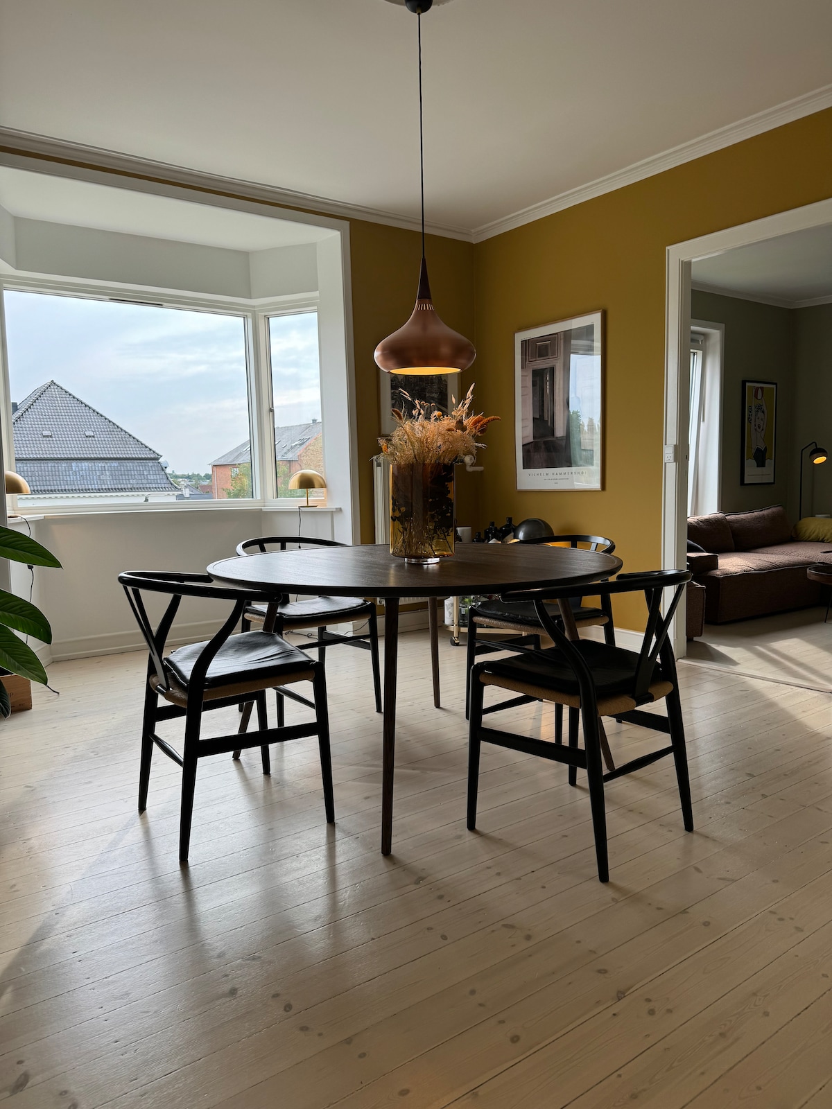 A round dining table is accompanied by four black chairs, featuring a floral centerpiece. Large windows provide natural light and views of the surroundings. The room is painted in a warm yellow tone, enhancing the minimalist decor and inviting atmosphere.