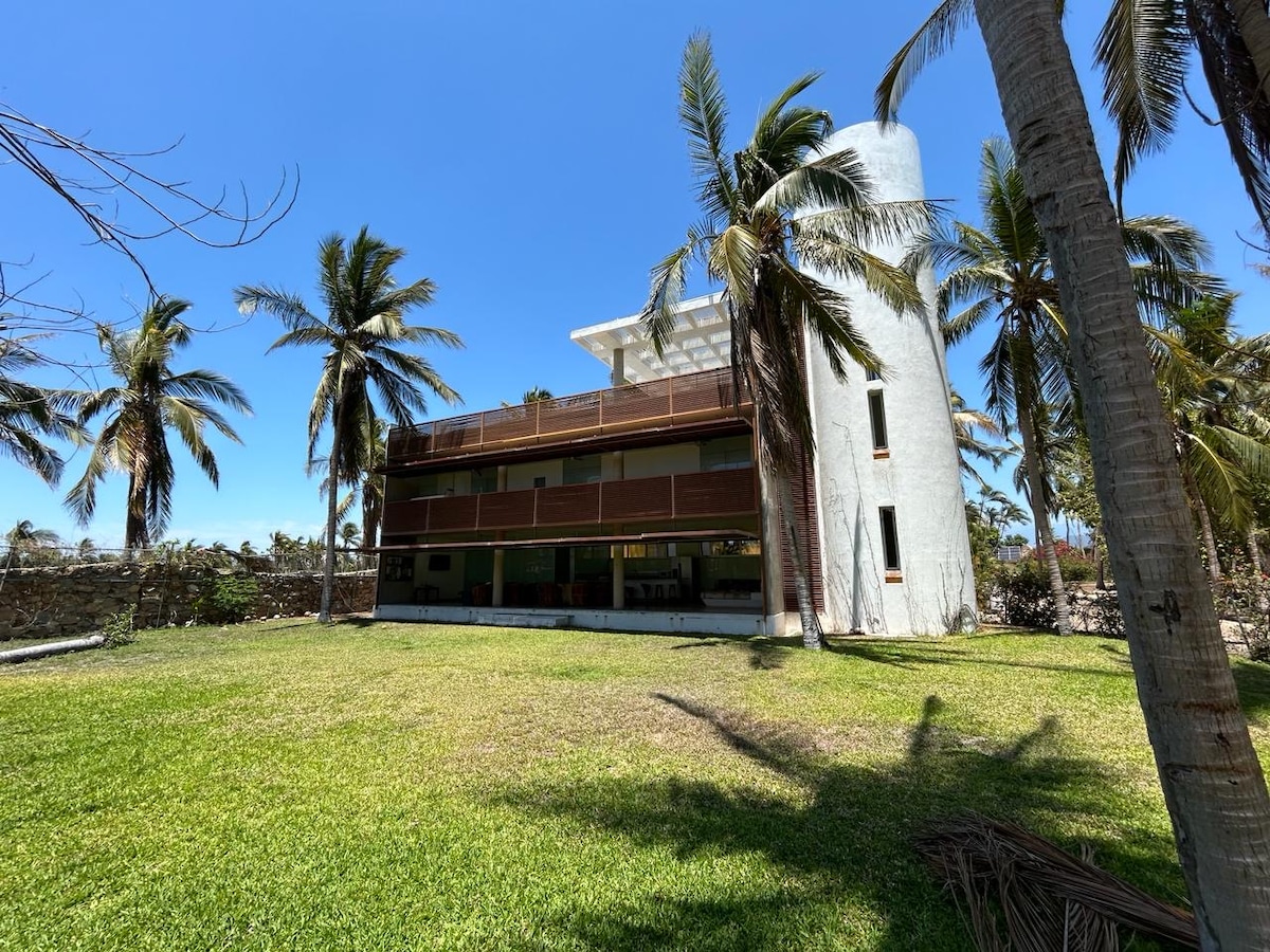 A spacious property is surrounded by well-maintained green grass and palm trees. The building features a multi-level design with a white exterior and wooden accents. Large windows provide ample natural light, and a wide balcony offers outdoor seating space, blending harmoniously with the outdoor landscape.