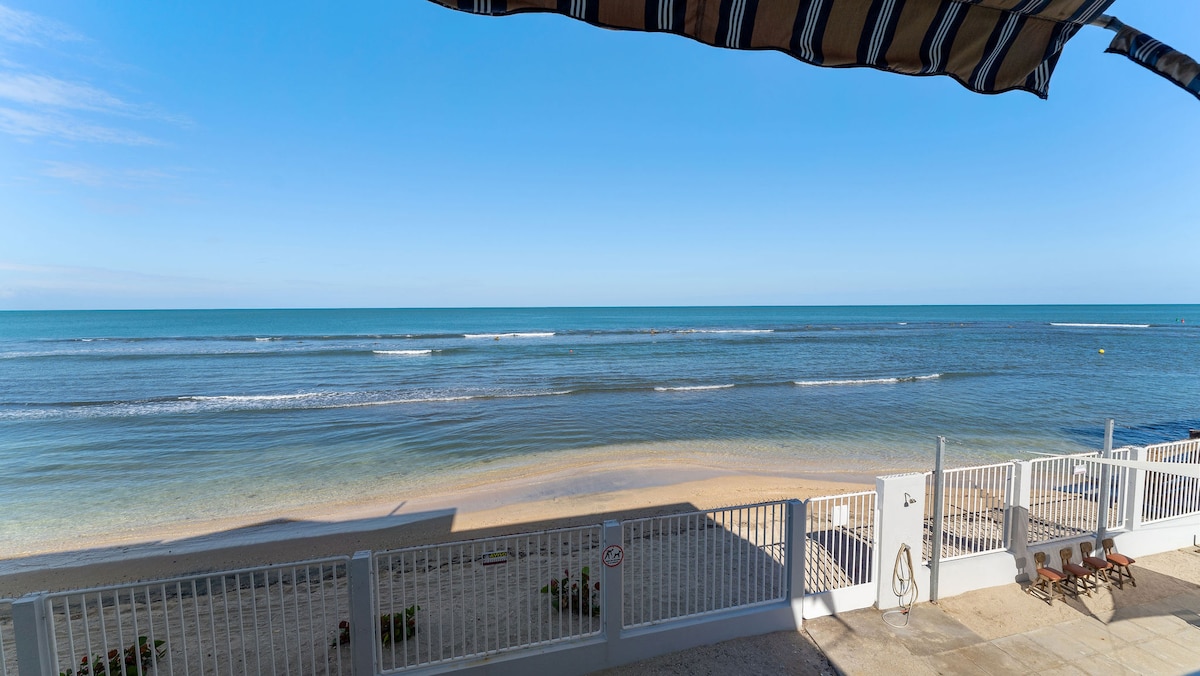 A serene beachfront view showcases calm, turquoise waters meeting a sandy shore under clear blue skies. Gentle waves create a soothing rhythm, while a striped awning provides shade over comfortable lounge chairs positioned along the pristine sand.