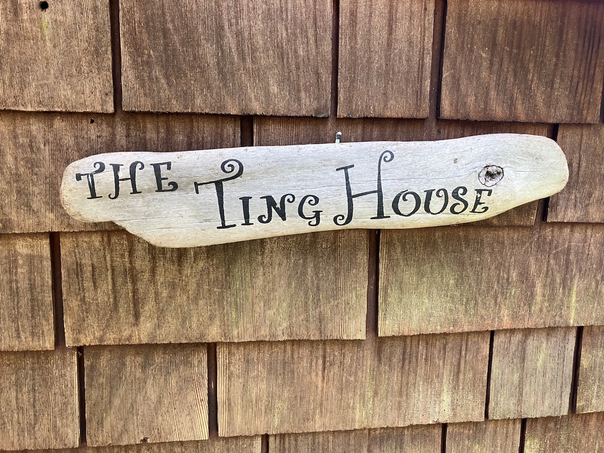 A weathered wooden sign hangs against a textured wall of cedar shingles, displaying the name 'THE TING HOUSE' in artistic lettering. The natural tones of the wood harmonize with the rustic charm of the cabin entrance.