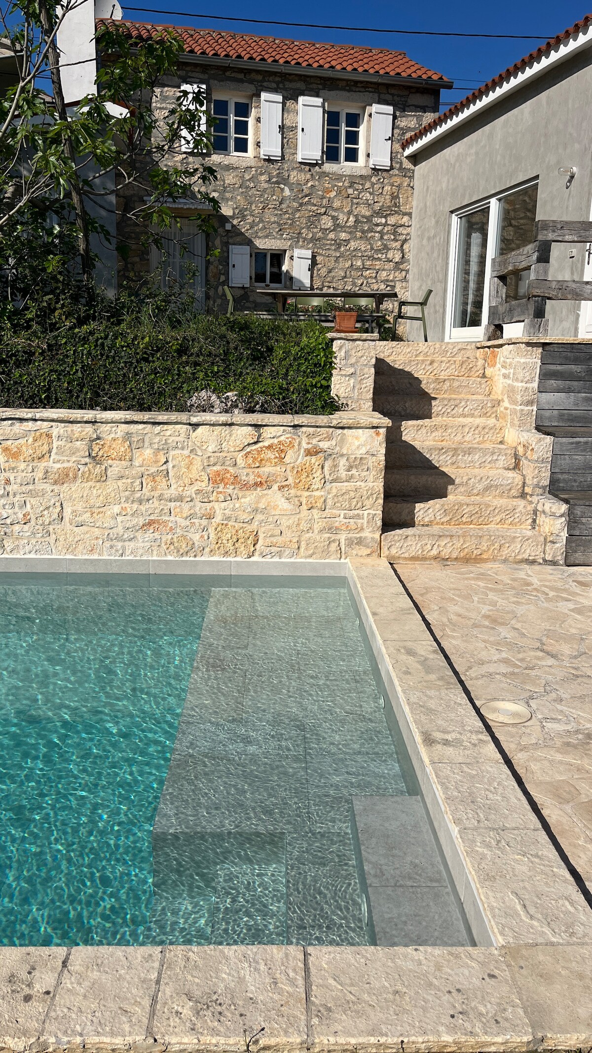 The image features a serene swimming pool with clear, inviting water, surrounded by natural stone paving. Steps lead up to a charming stone house with white shutters. Lush greenery frames the area, creating a peaceful outdoor setting.