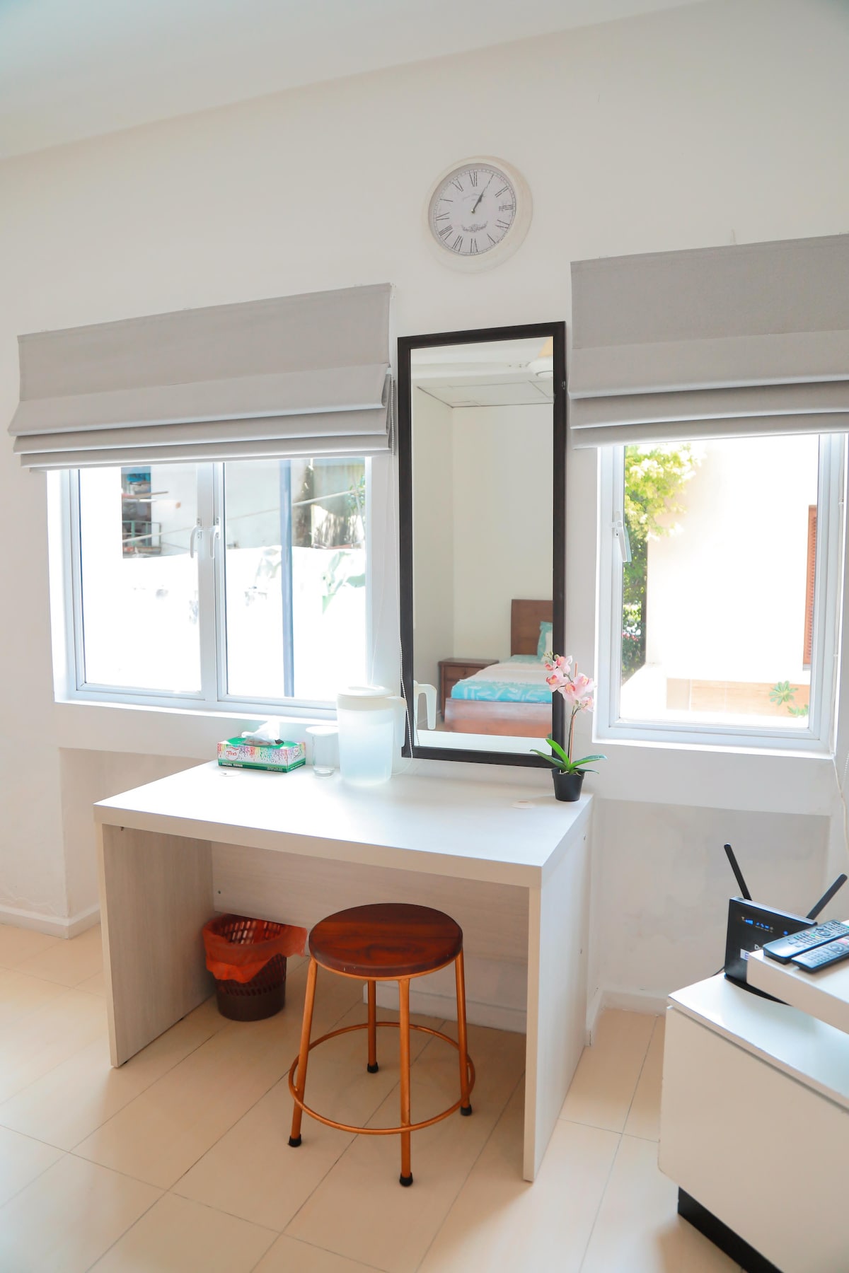 A bright area features a simple desk with a round wooden stool, complemented by a wall mirror. Natural light enters through two windows adorned with light gray shades. In the background, a softly made bed is observable, adding to the comfortable ambiance.