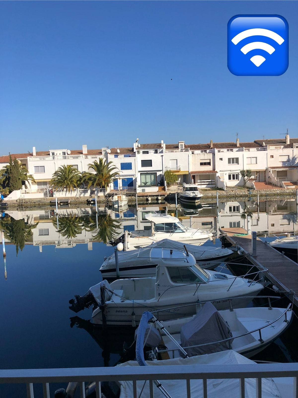 The image displays a serene view of a marina with several boats anchored on calm waters. The tranquil scene is punctuated by reflections of the surrounding buildings and palm trees, set against a clear blue sky.