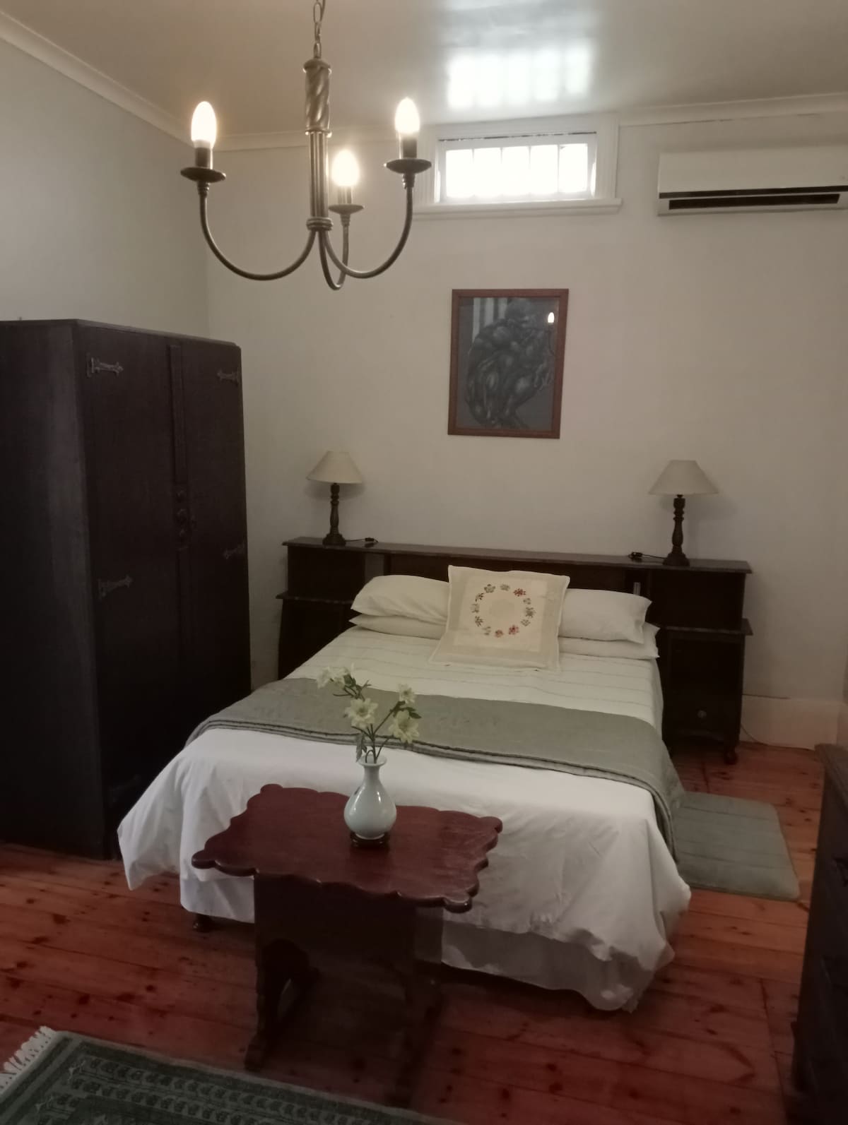 A cozy private bedroom features a spacious bed dressed in soft linens. Beside it, a vintage wooden nightstand holds a vase with fresh flowers. A large wardrobe stands against the wall, and a soft illumination is provided by a chandelier. An air conditioning unit is positioned above.