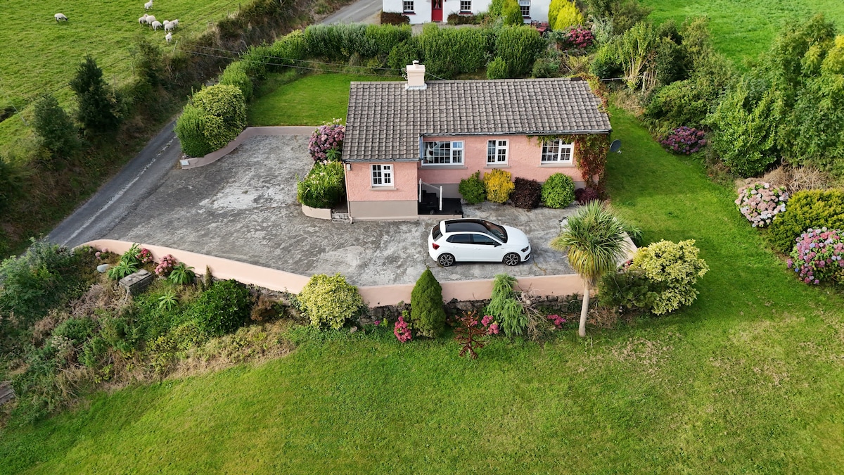 An aerial view of a cottage is presented, surrounded by lush greenery and blooming flowers. A compact driveway leads to the entrance, where a car is parked. The exterior features a light pink hue, complemented by a slate roof, creating a serene and inviting appearance.