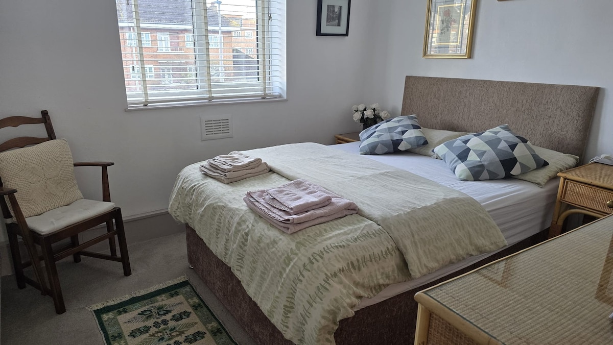 A comfortable bedroom features a spacious bed topped with patterned pillows and a soft blanket. A wooden chair is placed nearby, while neatly folded towels are arranged on the bed. Natural light filters in through the partially open window, illuminating the room.
