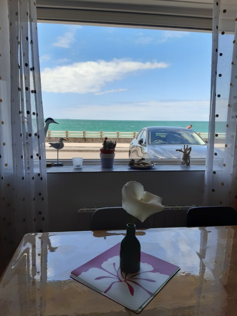 A view from a window showcases the sea, with gentle waves visible under a partly cloudy sky. A small table is displayed in the foreground, adorned with a flower in a glass vase, and decorative items positioned nearby.