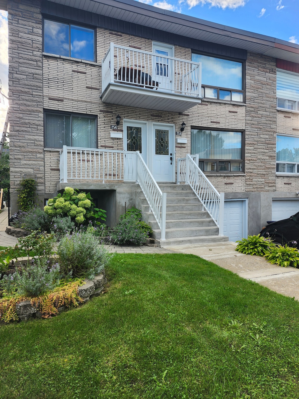 The exterior of the apartment building features a stone façade with white railings on the upper balcony. A well-maintained lawn with green shrubs and yellow flowers surrounds the concrete steps leading to the main entrance. Ample natural light is evident through the large windows.