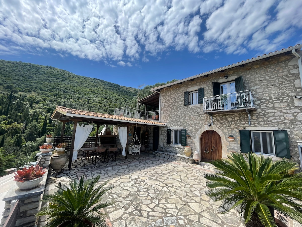 A traditional stone villa is featured with a spacious outdoor terrace adorned with greenery and rustic furniture. The structure combines stone and wood elements, showcasing an inviting entrance and balconies. The surrounding landscape includes lush hills under a blue sky with scattered clouds.