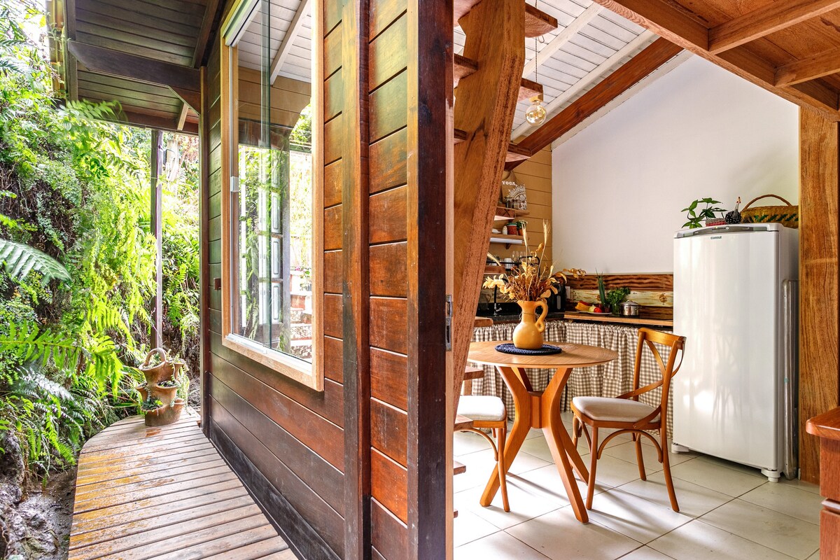 An inviting outdoor area features a wooden pathway leading to large windows, surrounded by lush greenery. The interior area showcases a cozy dining space with a circular table and chairs, complemented by a white refrigerator and wood accents in a well-lit environment.