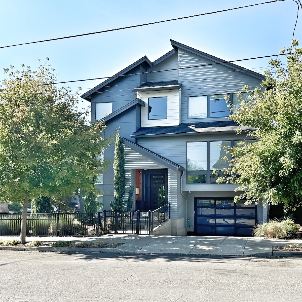 A modern two-story home is depicted, featuring a combination of gray and white siding. Large windows allow natural light to enter, while manicured shrubs and trees frame the entrance. A sleek garage door is visible, set against a clear blue sky.
